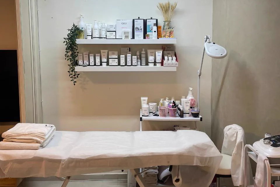 חדר הטיפולים בקליניקה של מיכל פאר: מיטת טיפולים לבנה עם מגבות מקופלות, מדפים לבנים עם מוצרי טיפוח, צמח ירוק, מנורת הגדלה ועגלת מכשור. אין אנשים בתמונה